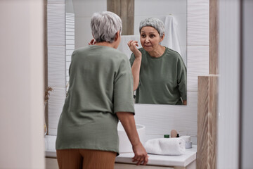 Back view of senior Asian woman looking in mirror and using facial cream