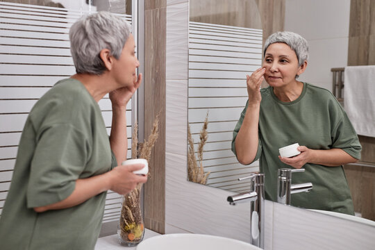 Side view portrait of senior Asian woman looking in mirror and using facial cream in anti aging routine