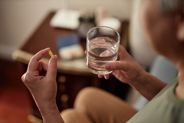 Close up of senior woman holding glass of water and taking pills at night, copy space