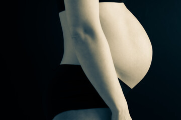 Side view of naked round belly of pregnant woman in underwear during last pregnancy month. Last month of pregnancy - week 36. Soft cropped shot. Black background. Partial toning.
