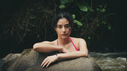 Beautiful Latina woman with curly hair, wearing a deep red swimsuit, leaning against a rock and...