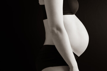 Side view of naked round belly of pregnant woman in underwear during last pregnancy month. Last month of pregnancy - week 36. Cropped. Black background. Sepia.