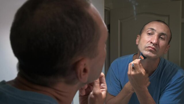 Daily hygiene of the male face. A forty-year-old man shaves his face with a razor while looking in the mirror.