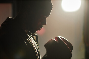 Profile view of young strong African American man in boxing gloves standing in front of camera against sunlit interior of sports club