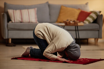 Young Muslim woman in grey hijab and casualwear bending forwards while standing on her knees on small red rug and saying prayers to Allah