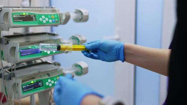Gloved hands holding the syringe and pushing its forcer. Medic puts the syringe attached to a wire on infusion pump machine.