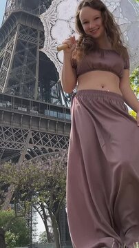 young beautiful girl in a blue dress sits near the Eiffel Tower near a green tree she straightens her braids looks around she is happy and very happy. High quality 4k footage