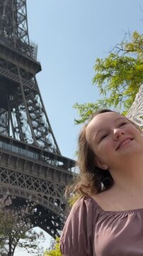 young beautiful girl in a blue dress sits near the Eiffel Tower near a green tree she straightens her braids looks around she is happy and very happy. High quality 4k footage