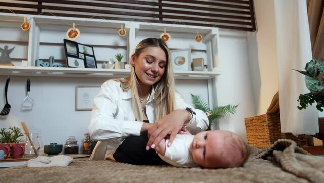 Beautiful smiling blonde woman putting on pants on her newborn boy. Mommy changing clothes to a baby. Low angle view.