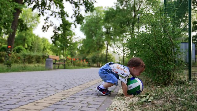 Toddler boy in jeans romper wants to lift the ball from the ground. Baby misses a little, nearly falls and looks at his palm.