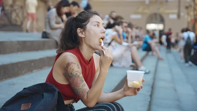 Portrait of young asian girl sitting on stairs and having lunch or snacking on fruit after walking in city. Woman sitting surrounded by other people. Journey to Florence, Tuscany. Tours to Italy.