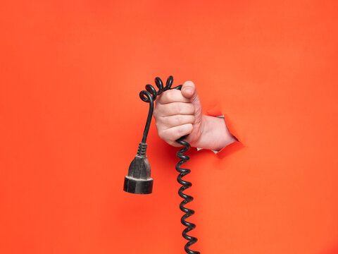 A man's hand holds a black spiral cable through torn orange paper. Concept, hand through torn orange paper.
