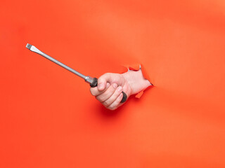 Hand holding a big screwdriver through torn orange paper. No face, concept.
