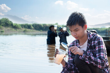 Asian boys hold magnifying glass, notebook, pen and transparent plastic tube which has clear water and cloudy water inside to compare and to do the water experiment in the river, nature study concept.