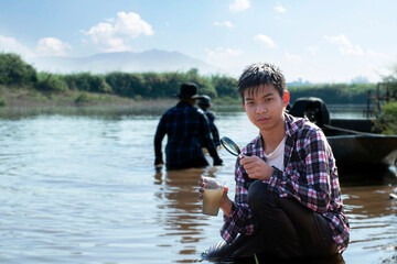 Asian boys hold magnifying glass, notebook, pen and transparent plastic tube which has clear water and cloudy water inside to compare and to do the water experiment in the river, nature study concept.