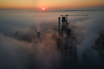 Obraz premium Aerial view of cement factory with high concrete plant structure and tower crane at industrial manufacturing site on foggy evening. Production and global industry concept