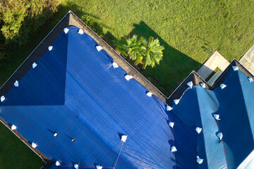 Aerial view of damaged in hurricane Ian house roof covered with blue protective tarp against rain water leaking until replacement of asphalt shingles - obrazy, fototapety, plakaty
