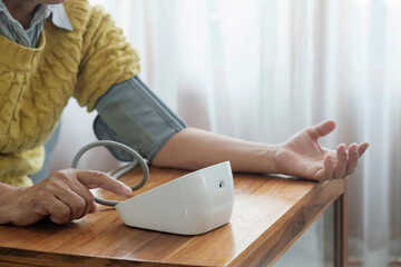 Asian Woman Measuring Arterial Blood Pressure Using Sphygmomanometer Cuff Sitting On Couch At Home.