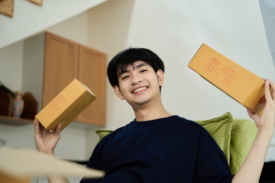 Young asian man unpacking  awaited parce looking inside, sitting at sofa, satisfied happy customer opening cardboard box with online store order, good shipping delivery service