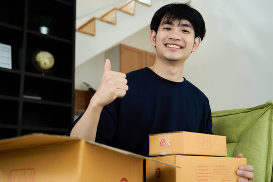 Young asian man unpacking  awaited parce looking inside, sitting at sofa, satisfied happy customer opening cardboard box with online store order, good shipping delivery service