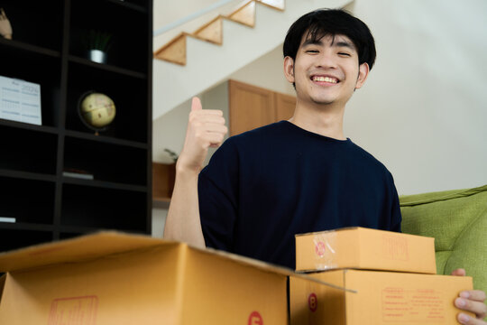 Young asian man unpacking  awaited parce looking inside, sitting at sofa, satisfied happy customer opening cardboard box with online store order, good shipping delivery service