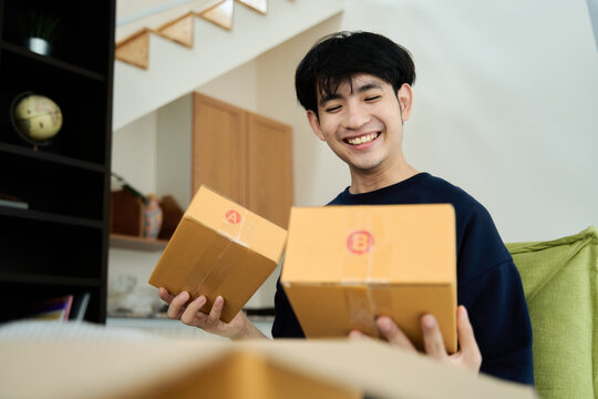 Young asian man unpacking  awaited parce looking inside, sitting at sofa, satisfied happy customer opening cardboard box with online store order, good shipping delivery service
