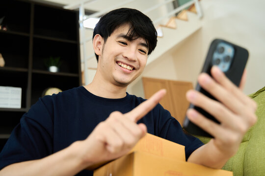 Young asian man unpacking  awaited parce looking inside, sitting at sofa, satisfied happy customer opening cardboard box with online store order, good shipping delivery service