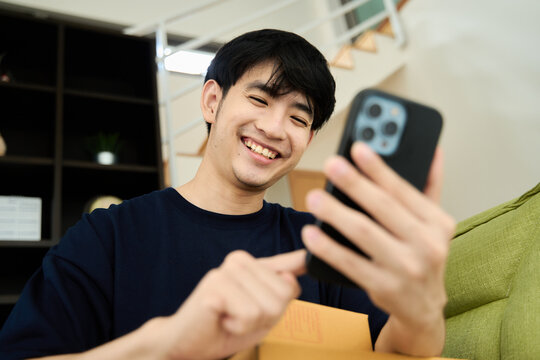 Young asian man unpacking  awaited parce looking inside, sitting at sofa, satisfied happy customer opening cardboard box with online store order, good shipping delivery service