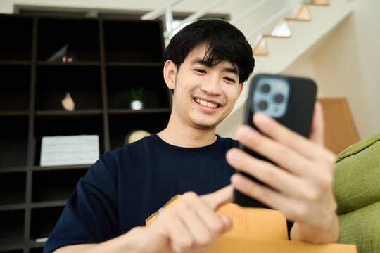 Young asian man unpacking  awaited parce looking inside, sitting at sofa, satisfied happy customer opening cardboard box with online store order, good shipping delivery service