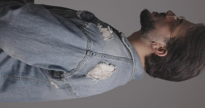 side view of casual bearded man with glasses wearing blue denim jacket and adjusting it while moving and looking forward in front of grey background