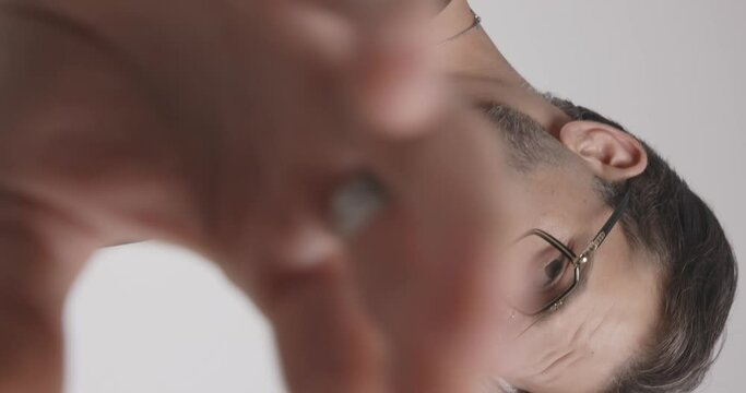 unshaved casual man with glasses adjusting necklace, moving in a side view pose before covering camera with his hand on light grey background