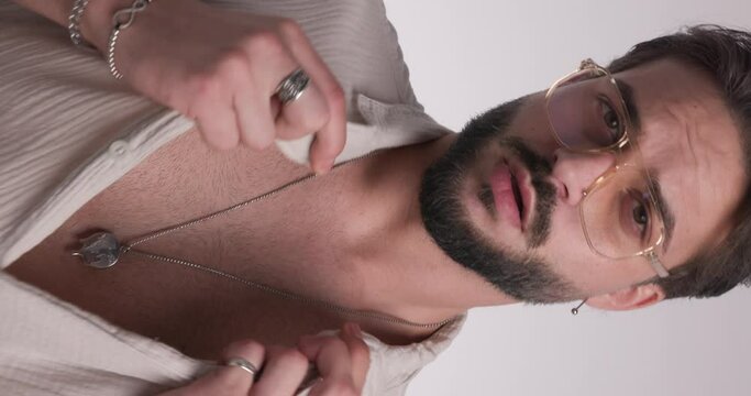 sensual young man holding hand to mouth and seductively touching lips, adjusting shirt and glasses while confidently posing in front of light grey background