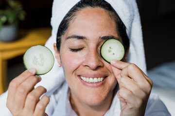 Latin adult woman applying facial mask on face with cucumber slices for exfoliation at home in...