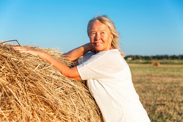 Naklejka na ściany i meble Smiling elderly woman in a white sweater and trousers in a field with hay rolls at sunset. Farming, freedom and nature. activity and longevity.