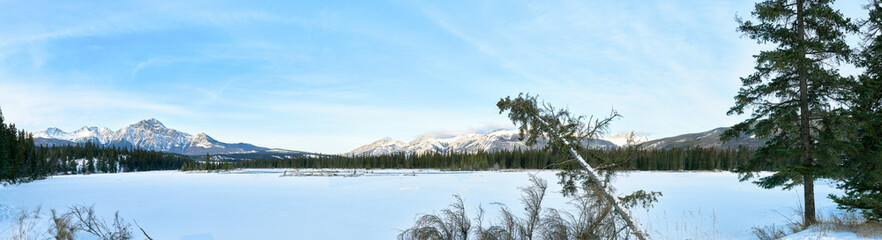 panoramic photo of the frozen lake of Jasper, Alberta, Canada.Rocky and snow-capped peaked mountains in the background. Green trees covered with snow in the foreground.
