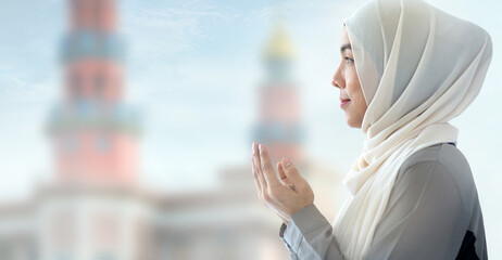 Side view of Muslim woman  in traditional clothes praying Dua with blurred mosque on background