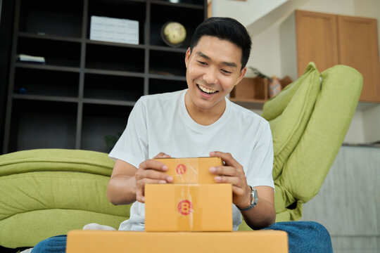 Young asian man unpacking  awaited parce looking inside, sitting at sofa, satisfied happy customer opening cardboard box with online store order, good shipping delivery service