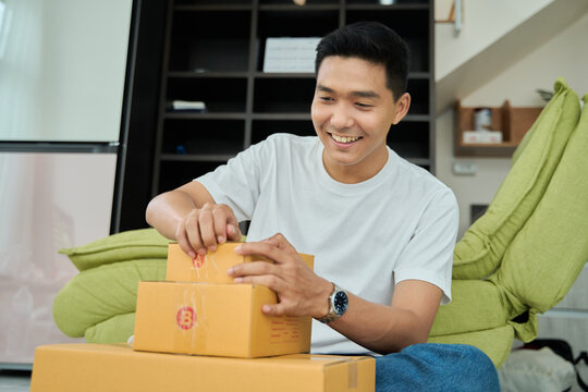 Young asian man unpacking  awaited parce looking inside, sitting at sofa, satisfied happy customer opening cardboard box with online store order, good shipping delivery service