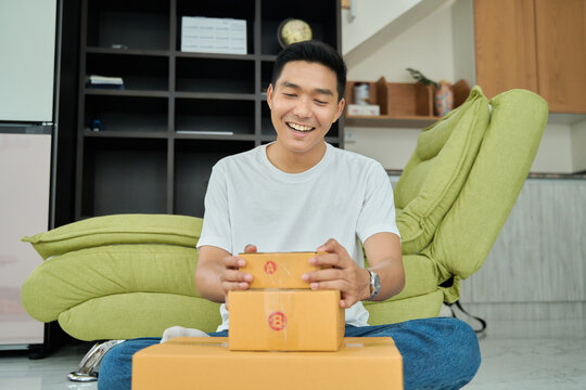 Young asian man unpacking  awaited parce looking inside, sitting at sofa, satisfied happy customer opening cardboard box with online store order, good shipping delivery service