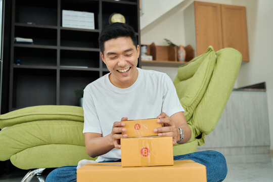 Young asian man unpacking  awaited parce looking inside, sitting at sofa, satisfied happy customer opening cardboard box with online store order, good shipping delivery service