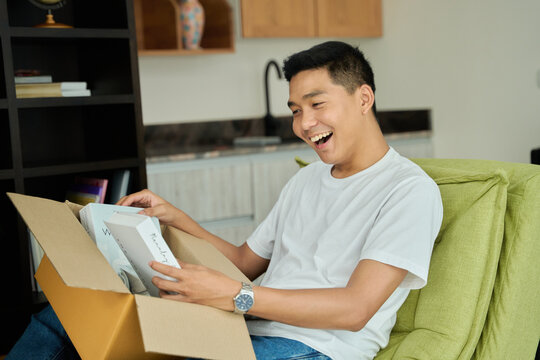 Young asian man unpacking  awaited parce looking inside, sitting at sofa, satisfied happy customer opening cardboard box with online store order, good shipping delivery service