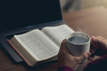 Hands holding a cup of coffee and a bible placed on a laptop were opened for learning to understand the Bible in order to pray to God and to ask God to protect himself and his family. Online bible.