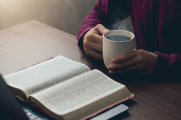 Hands holding a cup of coffee and a bible placed on a laptop were opened for learning to understand...