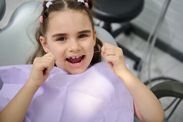 Close-up portrait of beautiful little girl using dental floss, brushing her teeth, sitting in the...