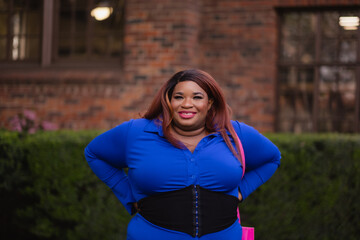 A woman poses on a college campus with her hands on her hips and a confident smile. She wears a blue dress, a hot pink purse, and bright makeup. Adding additional backstory, she is African American.