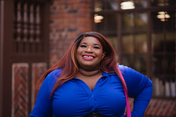 With a pink purse, a blue dress, and auburn hair, a college-age woman smiles joyfully in a campus-like setting. Her colorful fashion and African Americanness add backstory.