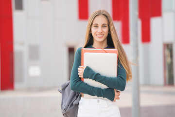 Teenage student holding laptop and notebooks, poses looking at camera standing in front of high...