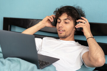 Young arab man using laptop wearing headphones watching film sitting in bed at home. Educational internet website advertisement and freelance lifestyle
