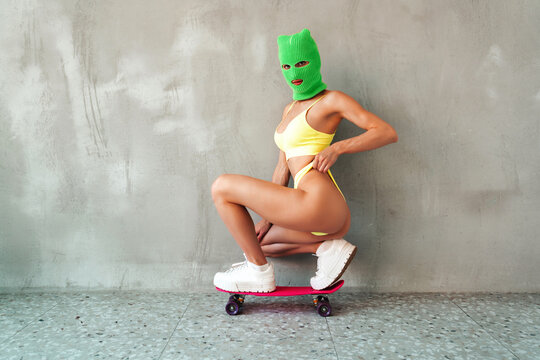 Beautiful sexy woman in green underwear. Model wearing bandit balaclava mask. Hot seductive female in nice lingerie posing near grey wall in studio. Crime and violence. Sits at penny skateboard