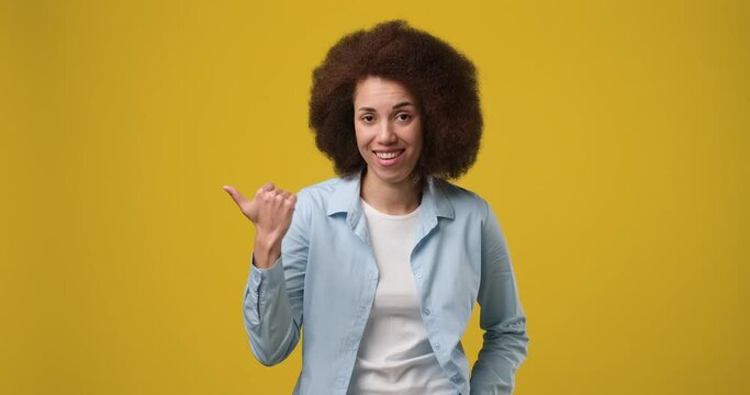 Cheerful expressive young african american woman in denim jacket white t-shirt pointing thumb aside on copy space workspace area isolated on orange studio background
